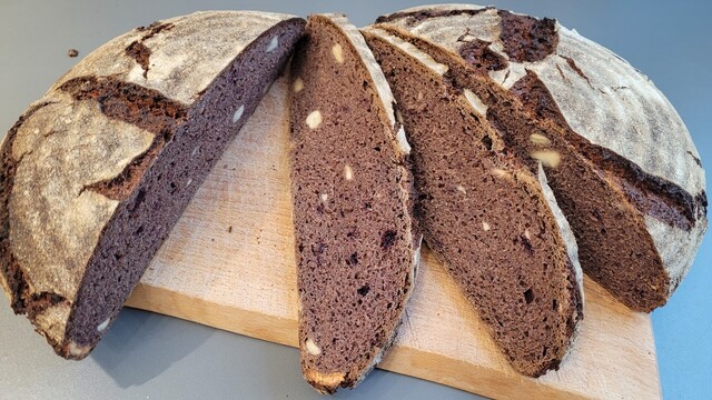 Derselbe Laib auf einem Schneidebrett, teilweise aufgeschnitten, so dass man die Krume sieht, die durch die Beeren blau-lila gefärbt ist. Auch die Cashewkerne sind zu sehen. 