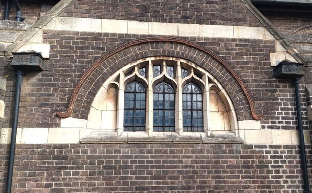 The East Wall of St Hilda's Church in Crofton Park, made of sober brick enlivened with horizontal lintels of stone, features an ornamental, semi-circular window. To either side of the window are two black drainpipes, each surmounted with a hopper boldly embossed with "1908".