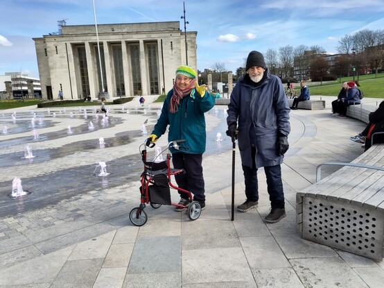 Two elderly people by town fountains smiling at camera