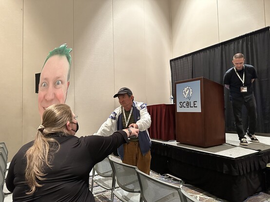 Katherine Druckman helps Shawn Powers - and his giant cardboard noggin - ask a question of Kyle Rankin. Shawn was watching the live stream from home, but was able to participate via phone! 