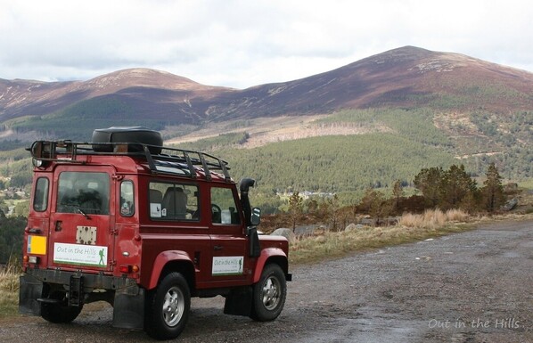 Red Landrover Defender showing Out In The Hills signage, parked by the side of the road, will a hillside in the background.