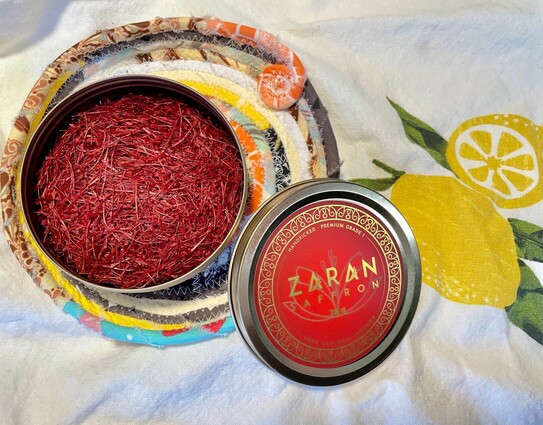 A round tin of saffron threads sitting on a dish towel with a print of a lemon. The saffron lid is adjacent to the tin and reads Zaran Saffron.