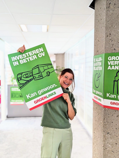 Ik sta in een gang met een poster van GroenLinks. Daarop staat “Investeren in beter OV. Kan gewoon. Met de afbeelding van een trein.  Ik draag een groene trui en lichtgroene broek.