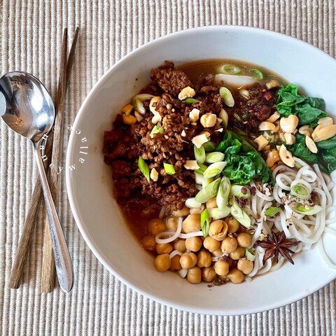 Overhead view of a bowl of Asian Soup called Chongqing,  with chopsticks and a spoon sitting on a placemat. The soup bowl contains spiced pork, baby bok choi, green onions, noodles, garbanzo beans, cashews, peppercorns, star anise, in a spicy broth.