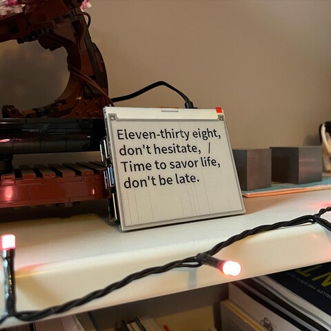 A small e-ink screen sitting on a bookshelf with a sentence displayed that reads: "Eleven-thirty eight, don't hesitate / Time to savor life, don't be late"