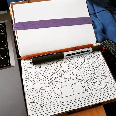Small sketchbook open with ink on paper art. A pen and pencil lie across the page. Outline drawing of a person as a traffic cone, their feet are the base, their legs the lower part, their torso the reflective stripe, their head the top tip of the cone. Behind is sky of circled stripes. Road streaks under the base.