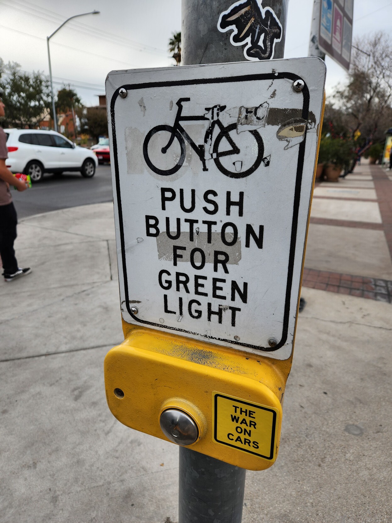 Bike level view of a silver beg button recessed in a yellow steel case. A square sticker for THE WAR ON CARS is mounted on the steel case. Above this is a sign with a depiction of a bike and the text PUSH BUTTON FOR GREEN LIGHT.