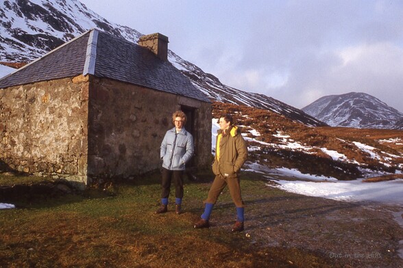 2 men stood in front of a mountain bothy, dressed for hill walking. Hills in the background with some snow covering.