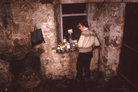 Man cooking on a window ledge in a mountain bothy. The interior of the building looks grim. Pots, pans, candles, and whisky bottles on the window ledge.