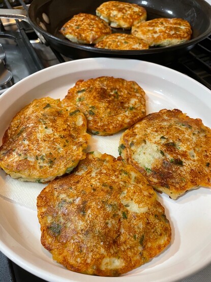A round white plate full of Irish potato pancakes, flecked with herbs, called Boxty. at the top of the photo, more pancakes are cooking on the griddle.