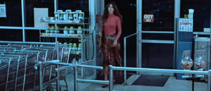 A woman in a red turtleneck and fringey 70s boho style skirt walks into a supermarket late at night. The parking lot was empty, but the supermarket....is not.