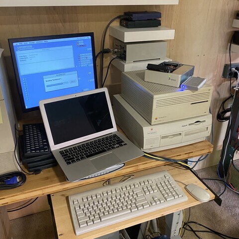 Workbench with a pair of beige Macintosh computers, a MacBook Air, external hard drives, keyboard, cables, and a flat screen monitor.