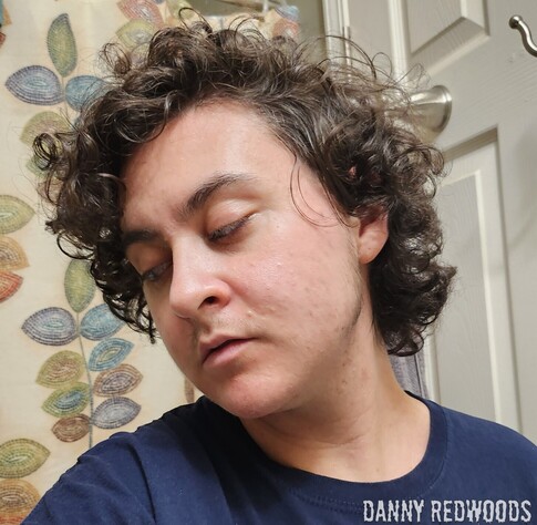 A young white trans man with messy curly brown hair looks down and to the side with parted lips and a very muted and almost bored look of pained and emotional yet peaceful acceptance and submission