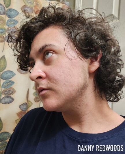 A young white trans man with messy curly brown hair has his chin tilted down but his blue/green/grey eyes look up and to the side, as though towards the Heavens. He has parted lips and a very muted and almost bored look of pained and emotional yet peaceful acceptance and submission