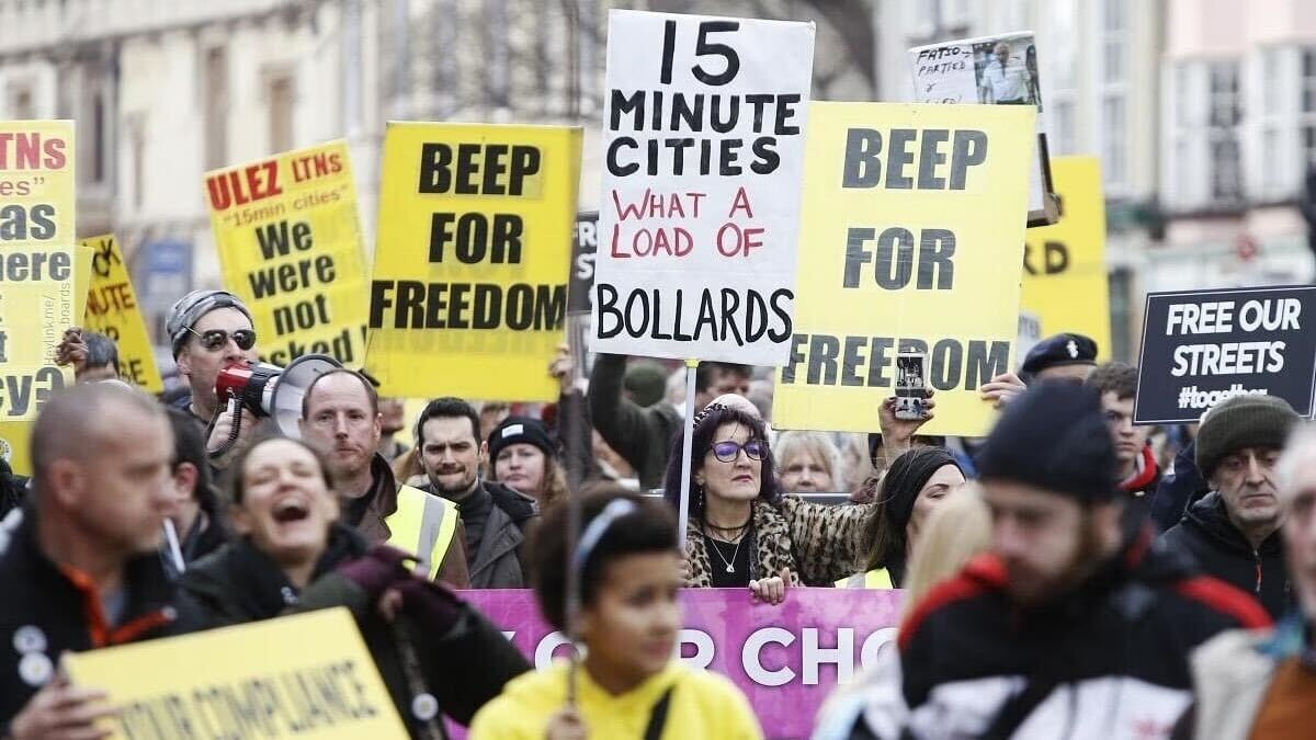 A protest against 15-minute cities. People with yellow and white signs saying "Beep for freedom" and "15 minute cities What a Load of Bollards."