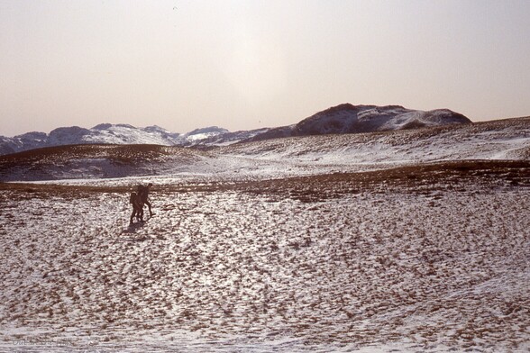 A snowy hillside, backlit by the sun, with 2 people walking across.