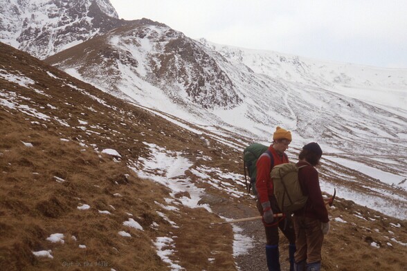A hillside, snowy higher up, two people in the foreground appear to be discussing something.