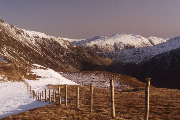 Looking towards the head of a valley, snow patches in the foreground and snow covered hills at the back. A series of wooden fence posts from top left to lower right.