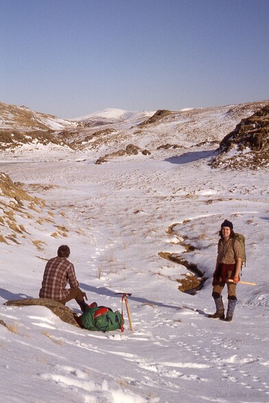 Looking up a snowy hillside, some rocks and grass showing through. One person sat looking away from the camera, another stood looking towards the camera.