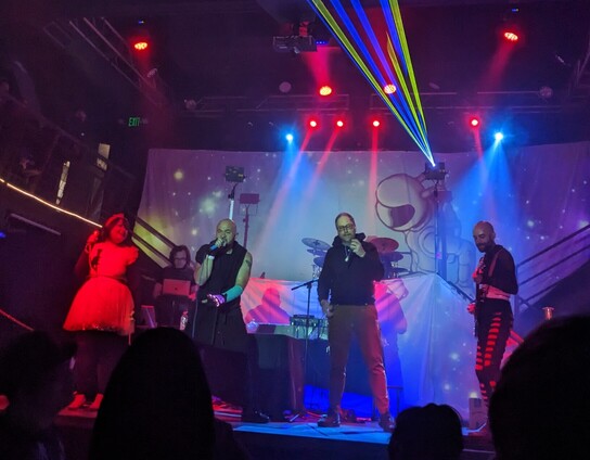 A photo of 5 performers on a stage. The lighting is filled with red and blue flood lights and lasers shooting overhead. 