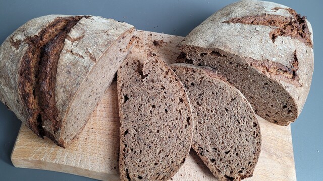 Ein ovaler Laib Brot mit bemehlter Kruste auf einem Schneidebrett, teilweise aufgeschnitten. Man sieht einen starken Riss an der Seite, die Krume ist mittelporig und einigermaßen gleichmäßig, man sieht darin auch ein paar Leinsamenkörner. 