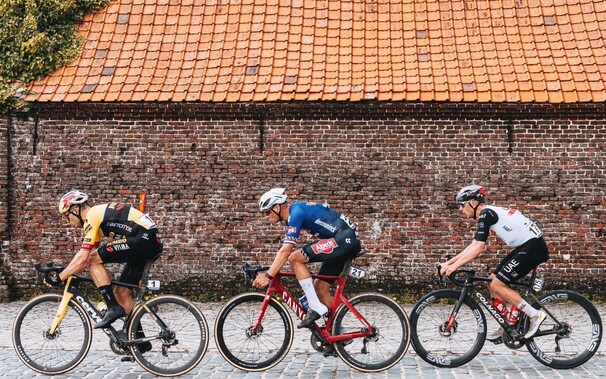 Cyclists Wout van Aert, Mathieu van der Poel and Tadej Pogacar ride from right to left on cobbles in front of an old brick wall with a red tiled roof in the E3 Prijs Harelbeke on Fri 24 Mar 2023.