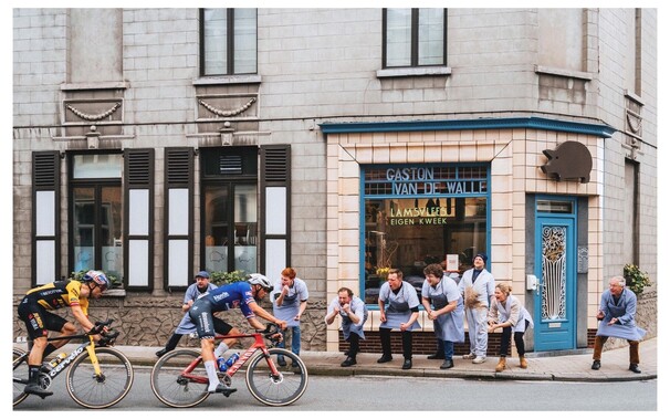 Cyclists Mathieu van der Poel and Wout van Aert ride from left to right around a corner in front of the shop window of butcher "Gaston Van De Walle" in Kluisbergen during the E3 Prijs Harelbeke on Fri 24 Mar 2023. All the butchers are outside cheering fanatically on the riders.