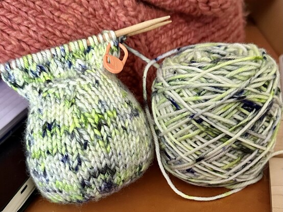 A small knitted object in variegated yarn - white, multiple shades of green, and a navy blue color. There is also the would yarn cake in the same yarn next to the small knitted object.