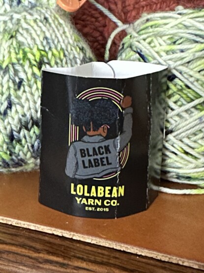 A yarn label for Lolabean Yarn Co. Black Label in front of a small knitted object and a wound yarn cake.