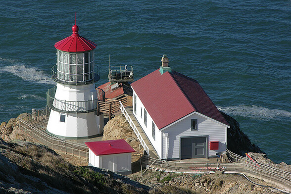 A squat, whitewalled lighthouse with a bright red roof and a whitewalled rectangular building next to it with a dark red roof on a rocky point overlooking dark blue Pacific water. There is a small lookout platform at the very edge of the point with a metal railing.