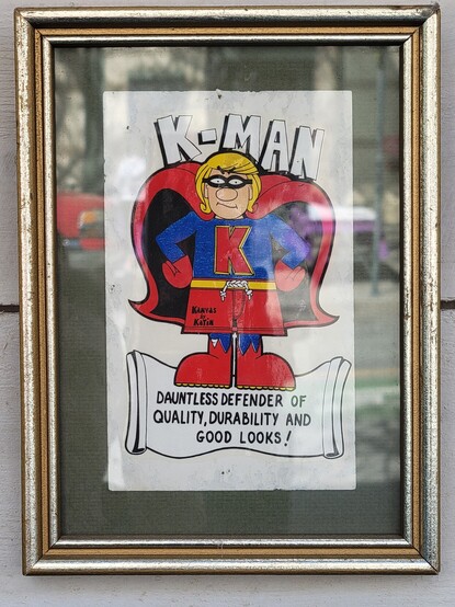 Drawing of a caped superhero. The top text reads K-Man and the bottom text on what looks like an unfurled banner reads "Dauntless defender of quality, durability, and good looks!" The inscription "Kanvas by Katin" appears on the character's shorts. The hero wears black glasses or goggles, a red cape, a blue long-sleeved shirt with a large letter K, red gloves, red shorts with a yellow waist band  tightened with a white cord, possibly blue tights, and red shoes with yellow soles. He has a large round nose and medium blonde hair. He rests his hands on his hips and has what looks like a serious expression. The drawing appears in a gold frame with green matting.