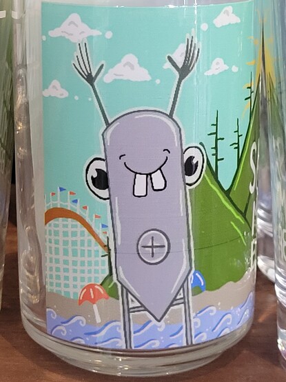 Cartoonish, anthropomorphic, smiling fermentation tank, with googly eyes, two buck teeth, and what look like hands raised above its head. It has what seems like a removable plug near it's midsection. It stands in front of the Santa Cruz Boardwalk roller coaster and mountains