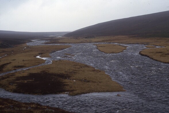 A river meandering and braiding over a wide valley which rises slightly to low hills. A ruined stone building on the far bank.