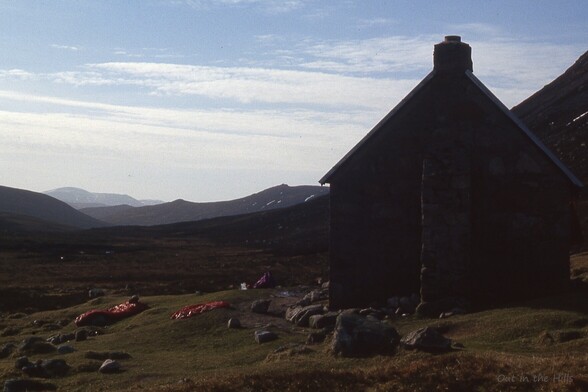 A stone building on a slightly sloping hillside, steeper hills to the side. The grassy patch immediately outside the building has some sleeping bags spread out in the sunshine.