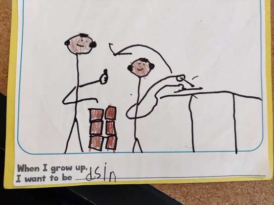 A picture of a worksheet. Two stick figures stand next to each other, in profile. One is shown drawing on a piece of paper. Another is standing next to a pile of what appear to be bricks, giving a thumbs up to the other. Underneath is a complete-the-sentence question: 'When I grow up I want to be...'. My kid has filled in the blank and written 'dsin'