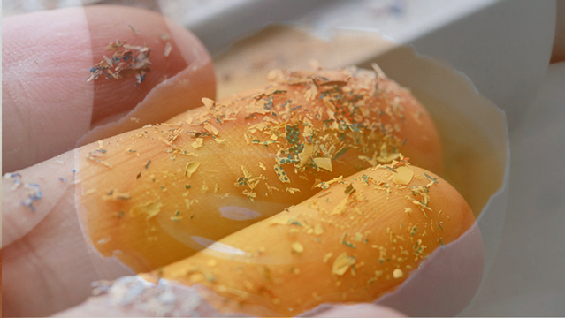 Photo of two human fingers covered in tiny pieces of plastic, superimposed on a photo of half a cracked eggshell. 