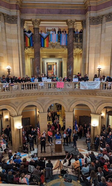 Photo of Trans Day of Visibility Festivities in the Minnesota State Capitol. Peggy Flanagan (Lt Gov) is speaking amongst a crowd in the Rotunda with trans flags and signs everywhere. 