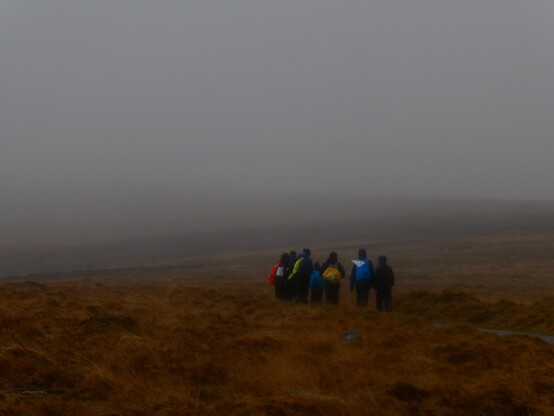 A group of young people in brightly coloured outdoor clothing with big rucksacks walking away from the camera across a wet and misty moorland.