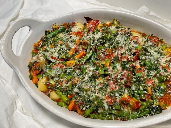 Fresh out of the oven, a white casserole dish filled with layers of colorful vegetables, toasted gnocchi, tomato sauce and topped with parsley and parmesan.