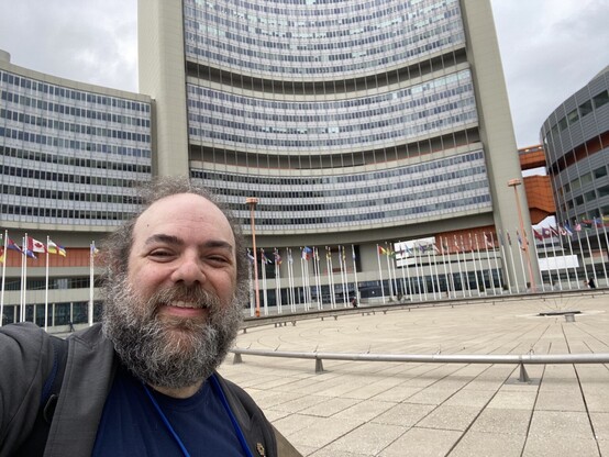 Andy standing in the circular central courtyard of the Vienna International Center, lined with flags. A several-story concrete building is on the circumference of the courtyard.