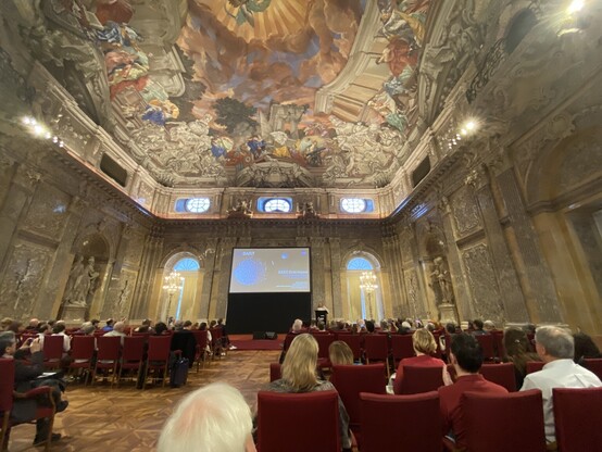 Nancy gives a presentation at the Austrian Academy of Sciences. The room has a painting on the ceiling that shows mythological figures. The walls have ornate, Baroque-era carvings.