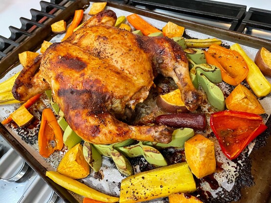 A fully roasted spatchcock chicken sitting on the roasting pan with assorted roasted vegetables: multi-colored carrots, bell peppers, and red garnet sweet potatoes. 