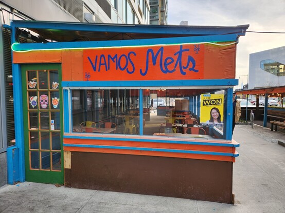 A colorful, enclosed wooden dining structure emblazoned with the words "vamos mets." In the background stands the architecturally significant Hunters Point South Library.
