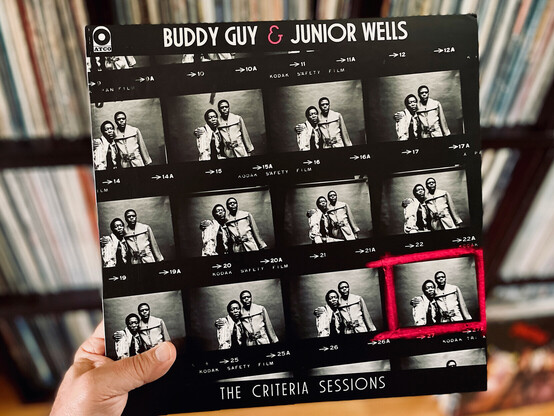 Hand holding a vinyl record (Buddy Guy's and Junior Wells' The Criteria Sessions) in front of a background of record shelves