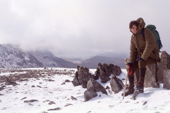 A flat bit of snowy ground with some rocks showing through. A person sat on the rocks, dressed for winter hillwalking. Hills at the back with their tops in thick cloud.