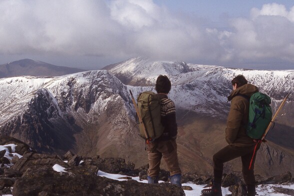 Two people dressed for winter hillwalking, with rucksacks and ice axes, looking away from the camera at the view. A range of hills, the upper parts covered with snow. Cloudy but bright.
