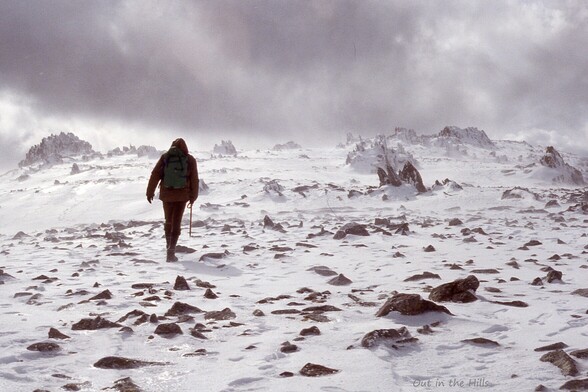 A snowy hillside with some boulders showing through. A person walking away from the camera, leaning into the wind. Cloudy.