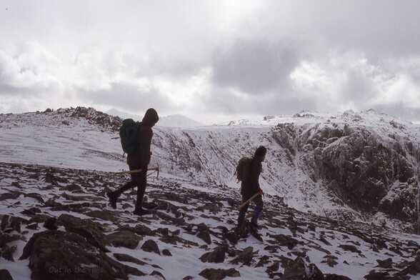 Two people walking down a slope on a hillside, boulders and some snow cover. Dull cloudy conditions.
