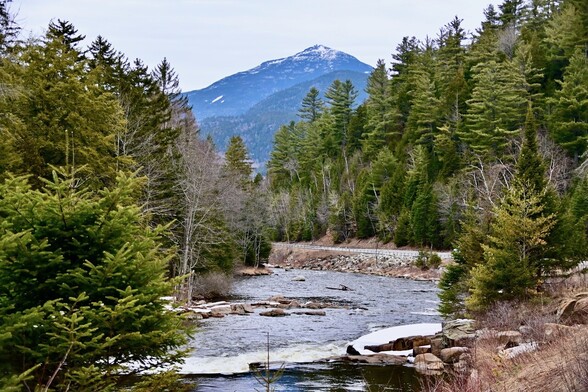 A mountain with some snow on the peak towers over a river surrounded by fir trees in winter. The river is fast flowing, and rushes over a small cascade in the foreground.