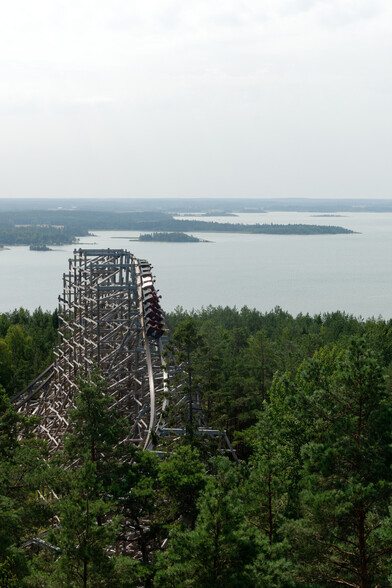 A train on Kolmården's RMC wooden coaster, Wildfire, enters an outward banked turn, high above the forest surrounding the ride. The body of water in the background is a massive sea inlet from the Baltic.
